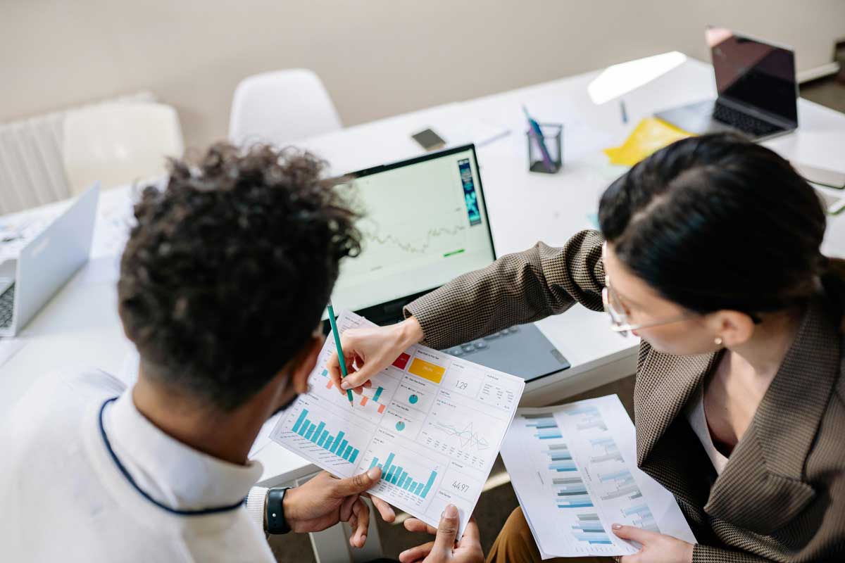 Two colleagues in a meeting room discussing financial charts and graphs on a laptop and paper