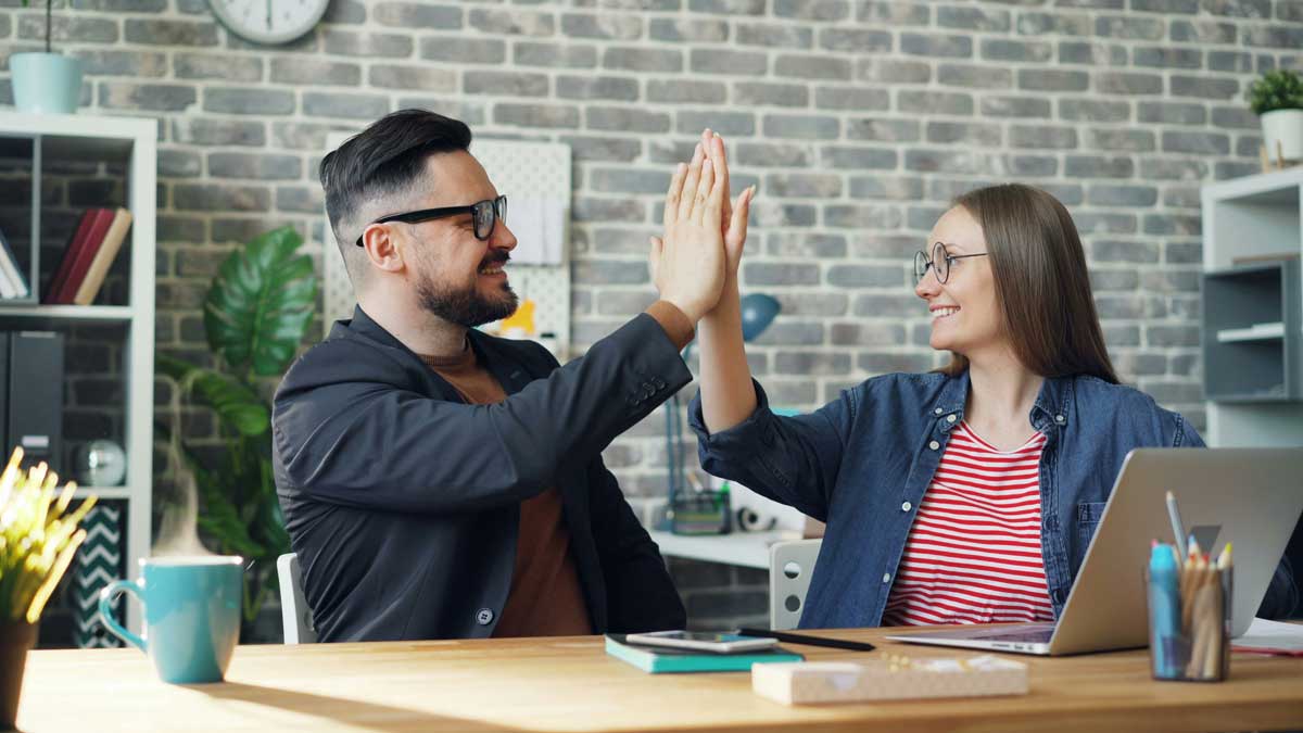 Happy colleagues man and woman doing high-five sitting at table in office while cup of hot tea is steaming on desk.