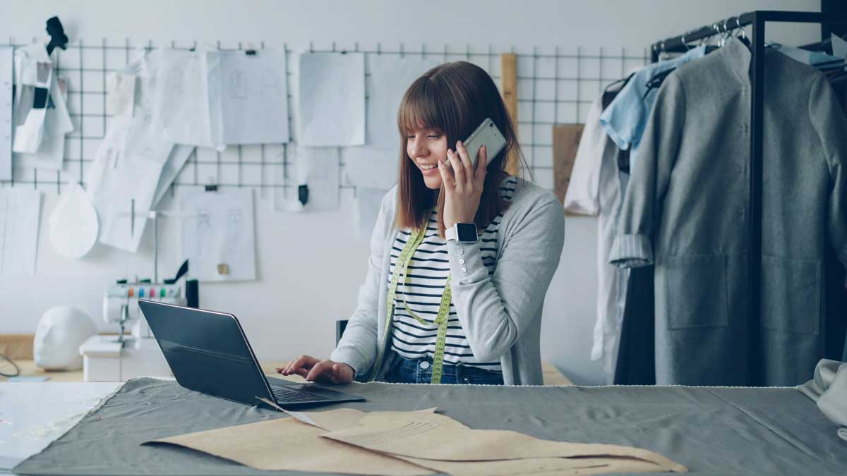 Small business owner is ordering fabric for her tailor's shop with mobile phone and working with laptop
