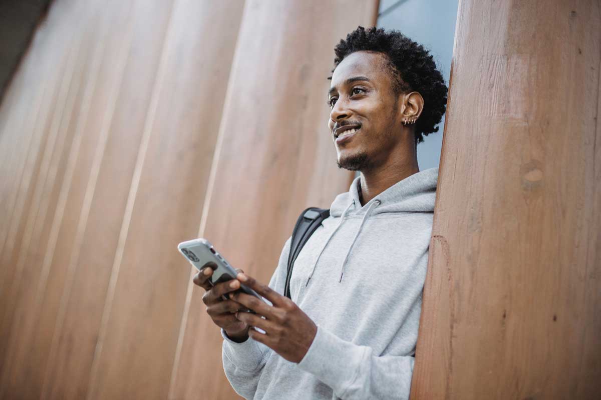 Smiling young man using smartphone outdoors, leaning on modern wooden architecture.