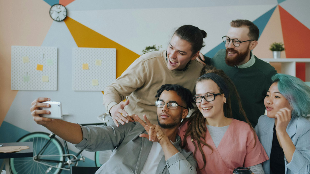 Joyful colleagues multi-ethnic group of young people are taking selfie with smartphone camera in office hugging gesturing having fun together