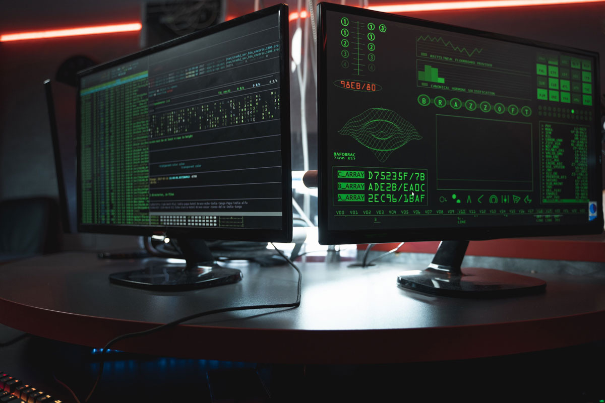 Close-up of dual computer monitors with green coding interfaces in a dark room, highlighting cyber security themes