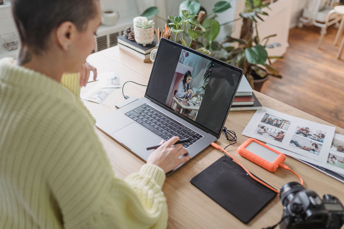 A woman edits photos on her laptop in a cozy home office, surrounded by plants and photography equipment