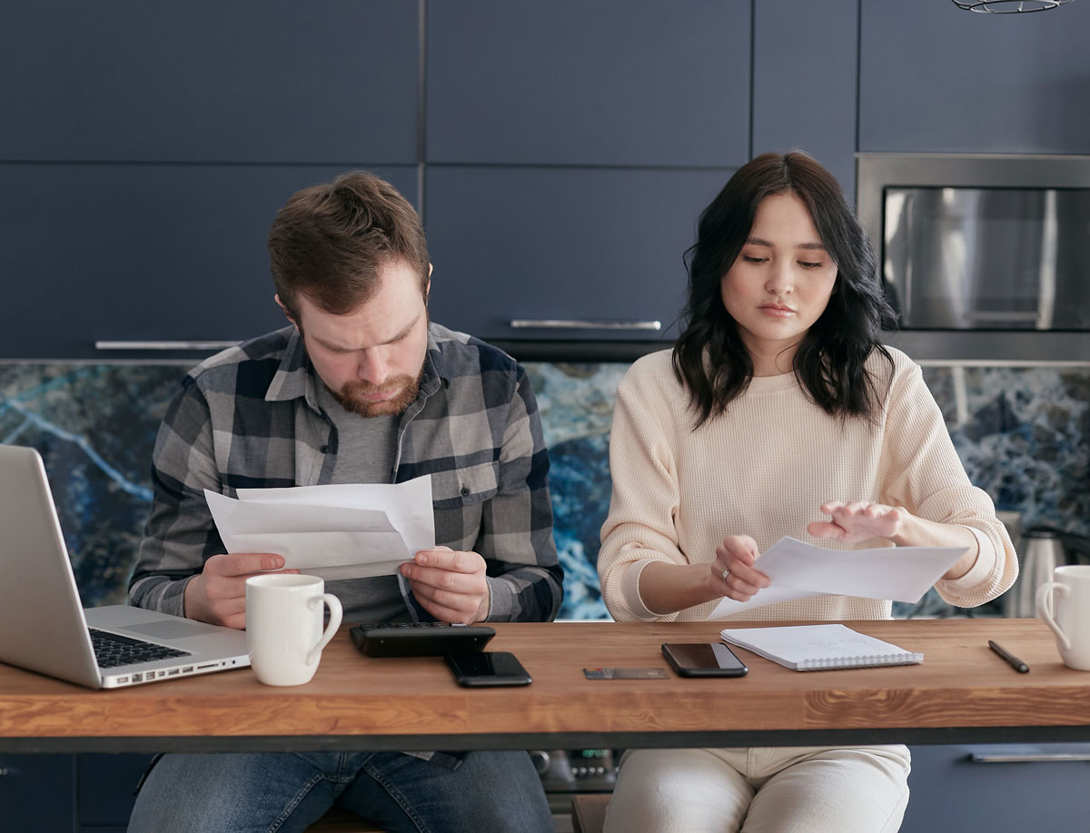 A couple reviews bills and manages finances on a kitchen table, looking concerned