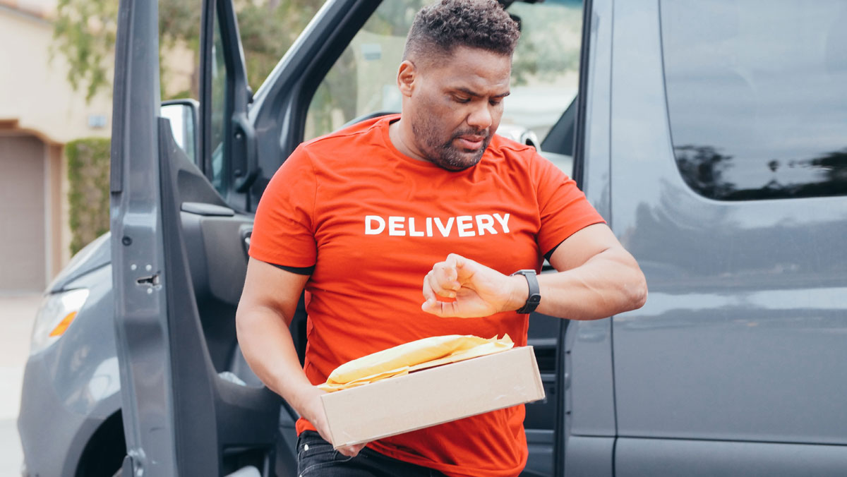 Delivery driver checks watch while carrying package from van outdoors.