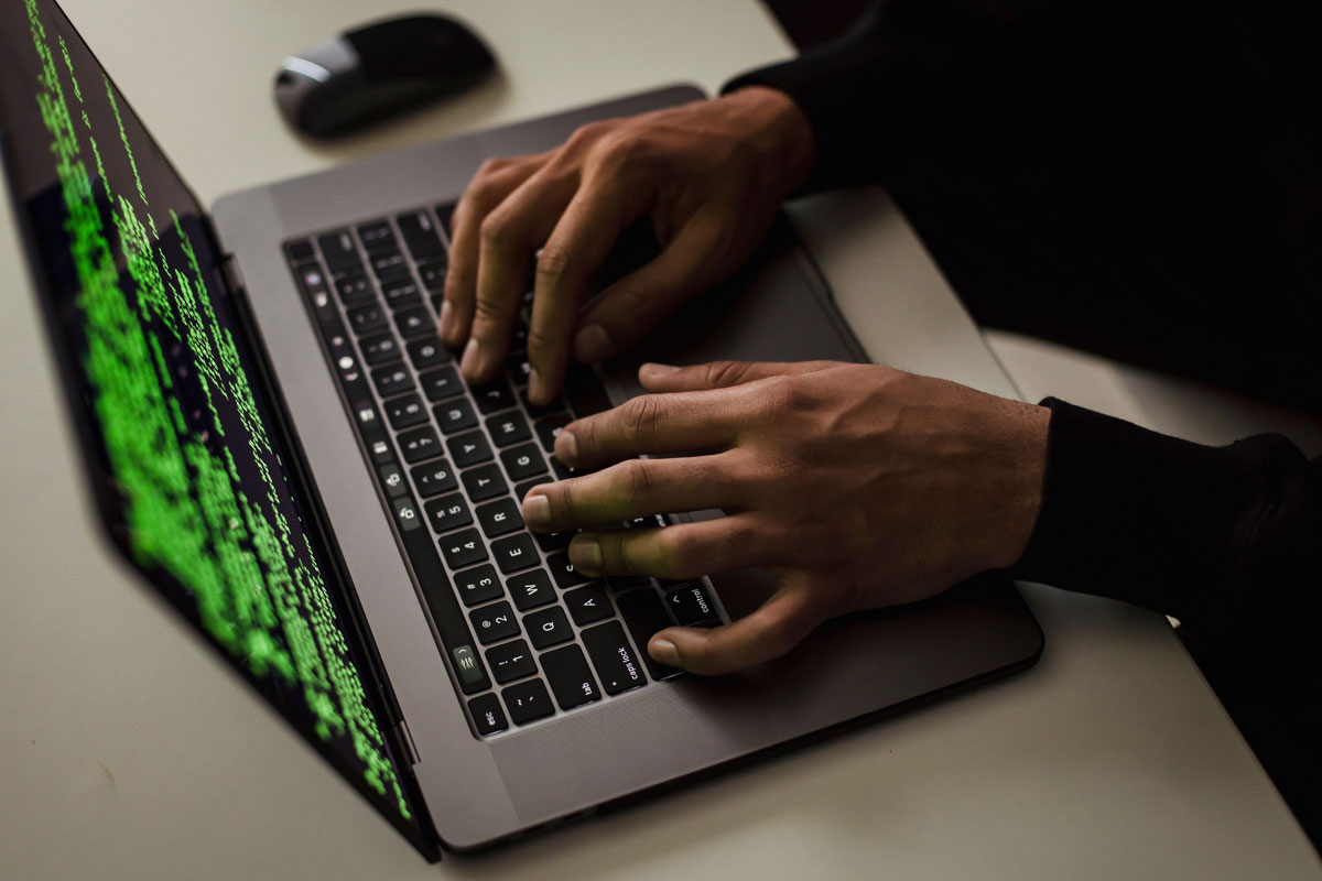 Close-up of hands typing code on a laptop with green text on screen, suggesting hacking or programming.