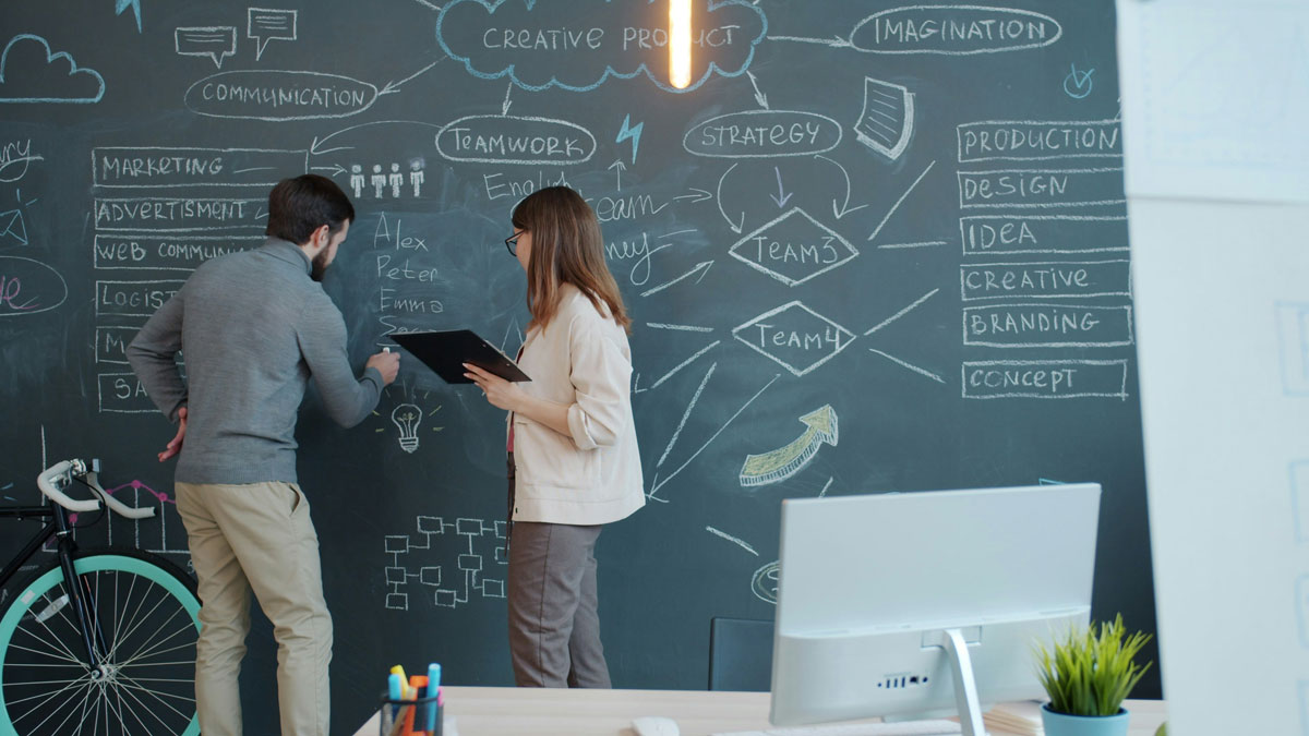 Businesswoman is talking to male colleague who is writing business information on chalkboard in office.