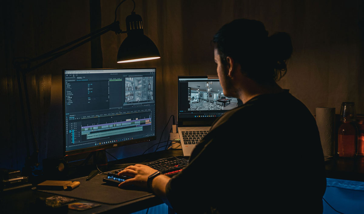 A man works at a computer editing video footage in a dimly lit workspace.
